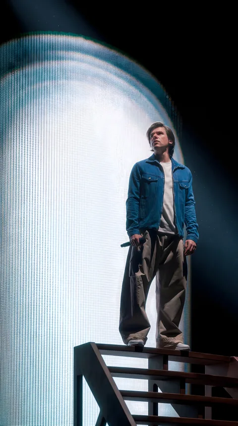 Orelsan en veste en jean debout sur un escalier devant un halo lumineux circulaire